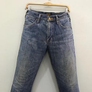 Op de afbeelding: Blauwe Wrangler-jeans hangend aan een houten hanger. De jeans hebben een klassiek ontwerp met vijf zakken, een knoop- en ritssluiting en het Wrangler-logo op de tailleband. De denim heeft een vervaagde, versleten uitstraling.