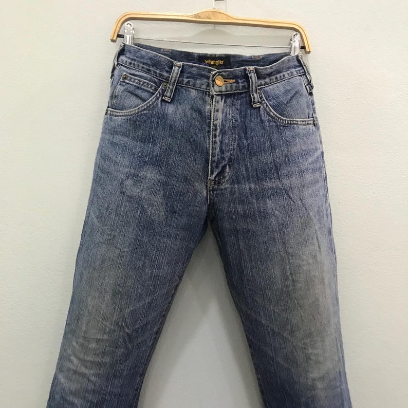 Op de afbeelding: Blauwe Wrangler-jeans hangend aan een houten hanger. De jeans hebben een klassiek ontwerp met vijf zakken, een knoop- en ritssluiting en het Wrangler-logo op de tailleband. De denim heeft een vervaagde, versleten uitstraling.