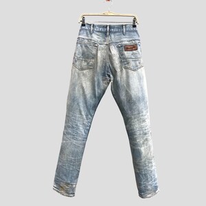 May include: Light blue denim jeans with a faded wash, hanging from a hanger. The jeans have a classic five-pocket design and a brown leather patch with the "Wrangler" logo on the back pocket. The jeans are a vintage style.