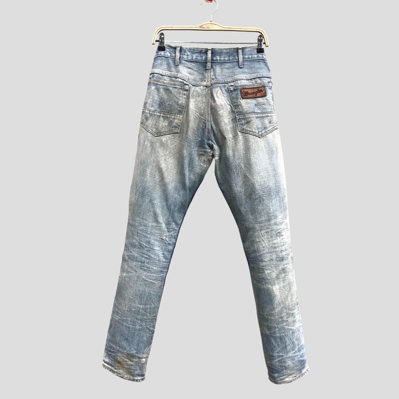 May include: Light blue denim jeans with a faded wash, hanging from a hanger. The jeans have a classic five-pocket design and a brown leather patch with the "Wrangler" logo on the back pocket. The jeans are a vintage style.
