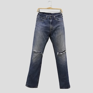 Op de afbeelding: Blauwe denim jeans met distressed details, waaronder gescheurde knieën. De jeans hangen aan een houten hanger tegen een lichtgrijze achtergrond. De jeans hebben een klassiek ontwerp met vijf zakken.