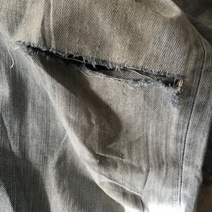 K&ouml;nnte beinhalten: Nahaufnahme von zerrissenem Denim-Stoff. Der hellblaue Denim zeigt einen zerrissenen Bereich mit ausgefransten Kanten, der die inneren Schichten des Stoffes freilegt. Die Textur des Denims ist sichtbar, mit subtilen Farbvariationen.