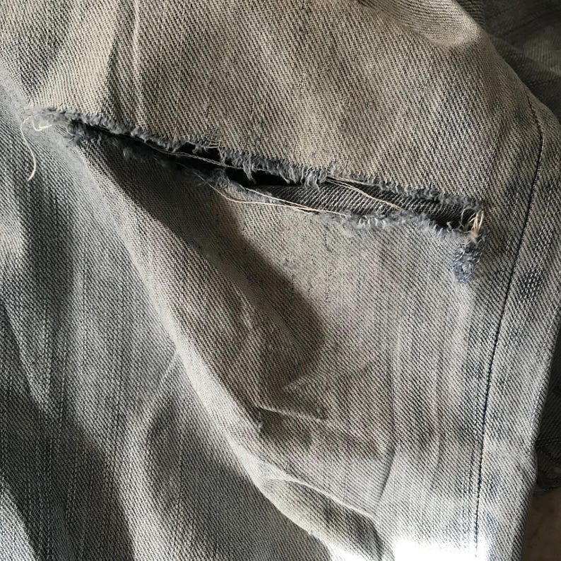 K&ouml;nnte beinhalten: Nahaufnahme von zerrissenem Denim-Stoff. Der hellblaue Denim zeigt einen zerrissenen Bereich mit ausgefransten Kanten, der die inneren Schichten des Stoffes freilegt. Die Textur des Denims ist sichtbar, mit subtilen Farbvariationen.