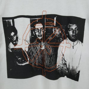 May include: Black and white photo of three people with an orange graphic overlay. The graphic is a stylized circle with lines radiating from the center. The people are wearing casual clothing. The photo is printed on a white t-shirt.