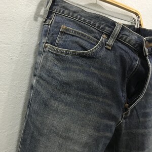 Pu&ograve; includere: Primo piano di jeans blu con cerniera parzialmente aperta. I jeans sono appesi a una gruccia di legno. Il denim ha un aspetto sbiadito e usurato, con dettagli delle tasche e cuciture visibili.