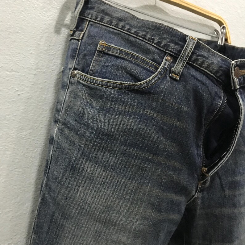 Pu&ograve; includere: Primo piano di jeans blu con cerniera parzialmente aperta. I jeans sono appesi a una gruccia di legno. Il denim ha un aspetto sbiadito e usurato, con dettagli delle tasche e cuciture visibili.