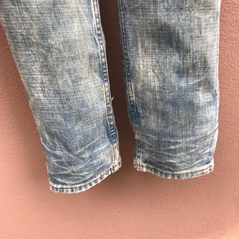 May include: Close-up of light blue denim jeans with distressed detailing. The jeans show signs of wear and tear, with faded areas and visible stitching. The bottom hems are slightly frayed, adding to the vintage aesthetic.