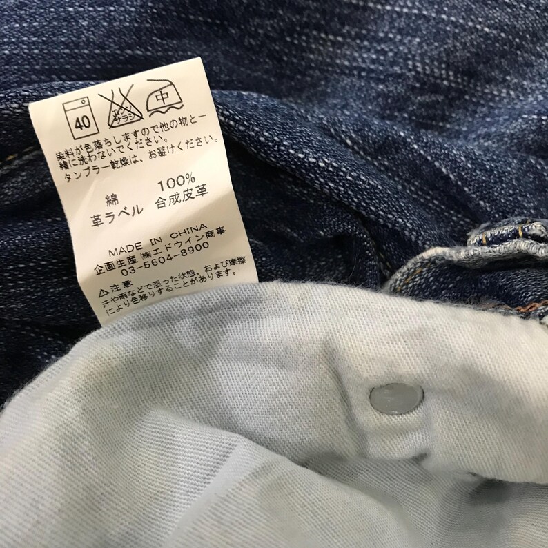 Op de afbeelding: Close-up van blauwe denimstof met een wit label. Het label heeft Japanse tekst en verzorgingssymbolen, waaronder een wasvoorschrift van 40 graden. Het label vermeldt ook dat de stof 100% katoen is en in China is gemaakt.