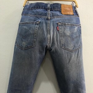 May include: Faded blue denim jeans with classic five-pocket styling. The back view shows the pockets, a leather patch, and a red tag. The jeans are hanging on a wooden hanger.