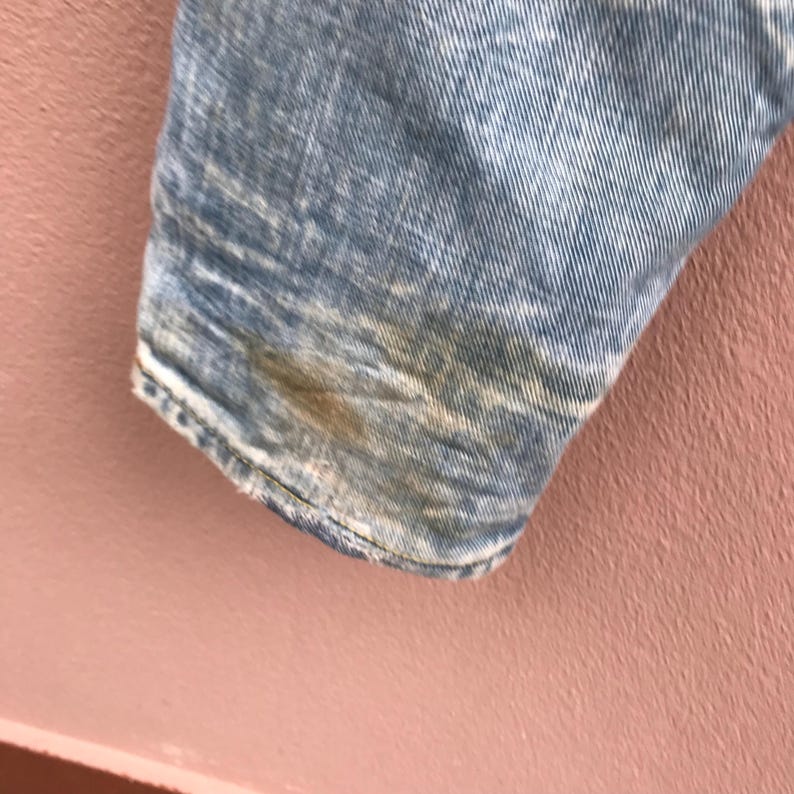 May include: Close-up of a light blue denim fabric with a distressed, acid wash effect. The fabric shows areas of discoloration and wear, with visible seams and a frayed edge. The denim has a vintage, worn appearance.