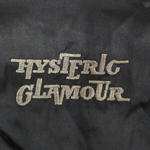 Może przedstawiać: Czarna naszywka haftowana z napisem "HYSTERIC GLAMOUR" w białym kolorze.