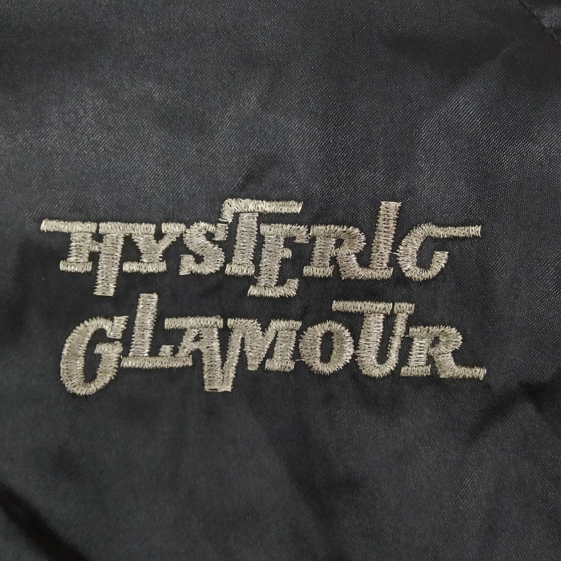 Może przedstawiać: Czarna naszywka haftowana z napisem "HYSTERIC GLAMOUR" w białym kolorze.