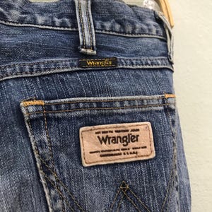 Op de afbeelding: Close-up van de achterkant van een blauwe Wrangler jeans. De jeans heeft een bruine leren patch met het Wrangler logo en tekst. De stiksels zijn geel en de jeans heeft een klassiek ontwerp met vijf zakken.
