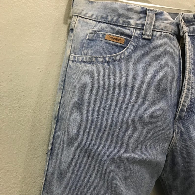 May include: Light blue denim jeans with a classic five-pocket design. The jeans feature a zipper fly and a small brown label with the "Wrangler" logo on the front pocket. The jeans are hanging on a hanger.