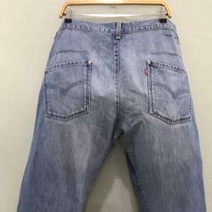 May include: Light blue denim jeans featuring a high-waisted design with two back pockets, each accented with a signature arcuate stitch pattern. A small red tab is visible on the right back pocket, and the jeans are suspended from a wooden hanger.