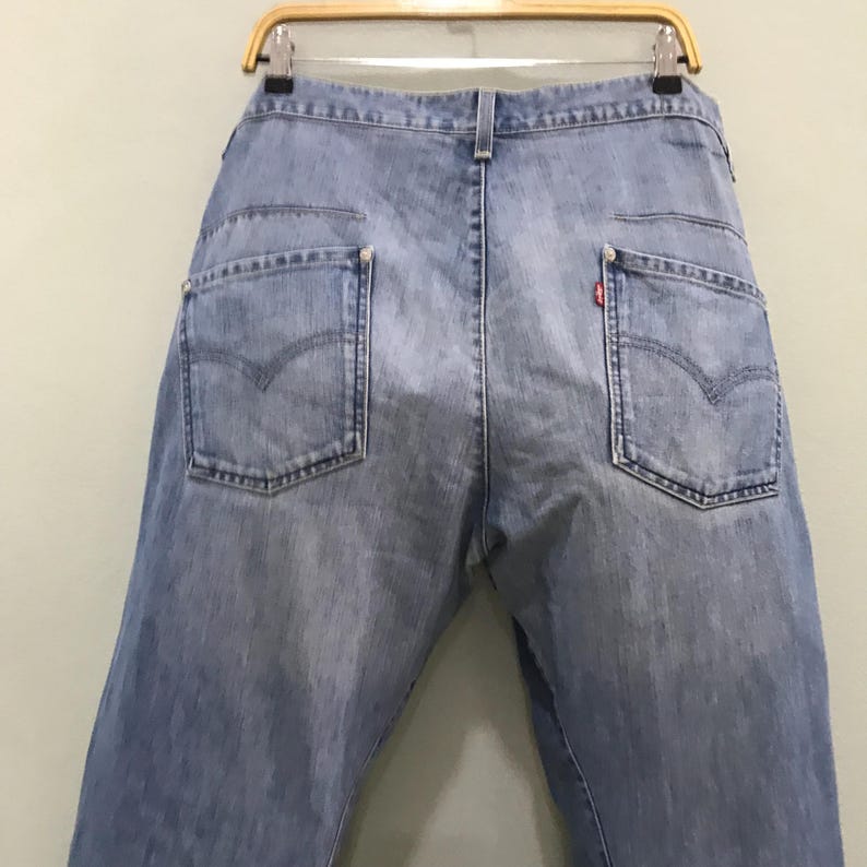 May include: Light blue denim jeans featuring a high-waisted design with two back pockets, each accented with a signature arcuate stitch pattern. A small red tab is visible on the right back pocket, and the jeans are suspended from a wooden hanger.