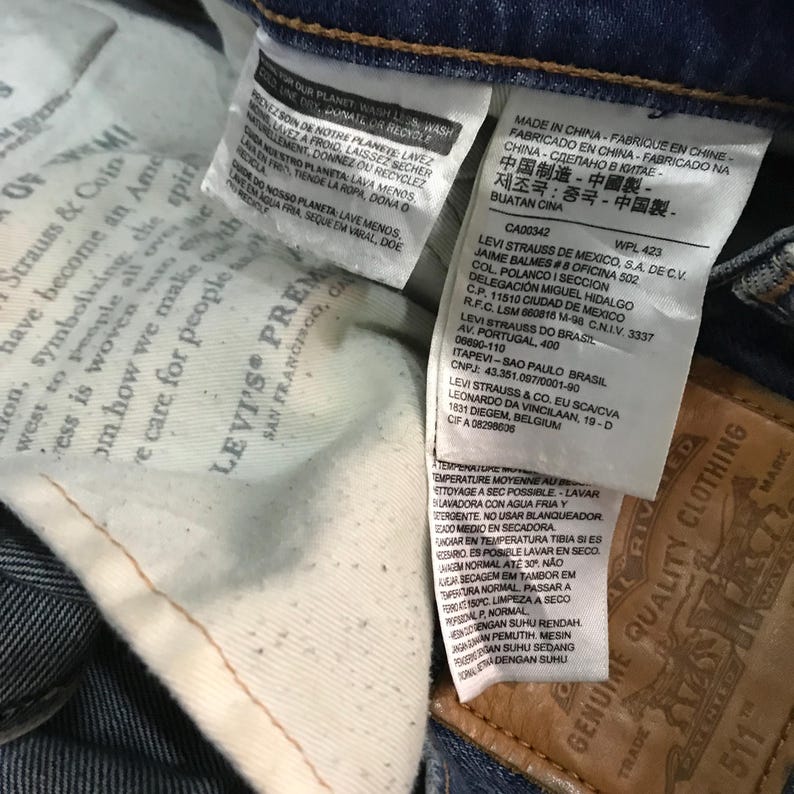 May include: Close-up of the inside of a pair of blue jeans, showing multiple white tags with printed text. One tag reads "LEVI'S PREMIUM SAN FRANCISCO, CA". Other tags include washing instructions and manufacturing information.