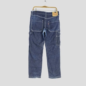 Puede incluir: Pantalones de trabajo de mezclilla azul oscuro con costuras blancas y múltiples bolsillos. Los pantalones están colgados de una percha de madera. Una etiqueta roja y beige de Levi's es visible en la parte posterior.