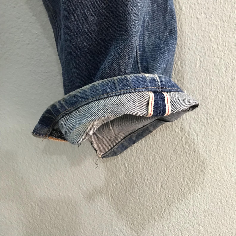 May include: Close-up of the cuffed hem of blue denim jeans. The cuff reveals a red and white stripe detail on the inside. The jeans are a medium wash and show signs of wear.