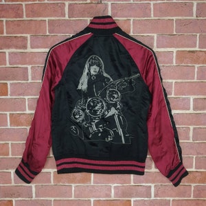 Może przedstawiać: Czarna satynowa kurtka bomber z czerwonymi rękawami i nadrukiem graficznym przedstawiającym kobietę jadącą na motocyklu. Kurtka ma żebrowany kołnierz, mankiety i d&oacute;ł.