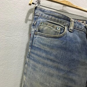 以下が含まれることがあります： 色あせたウォッシュ加工が施された、明るいブルーデニムのジーンズのクローズアップ。ジーンズは、小さなコインポケットを含む、クラシックな5ポケットデザインが特徴です。生地にはわずかな摩耗が見られ、側面に赤いタグが見えます。