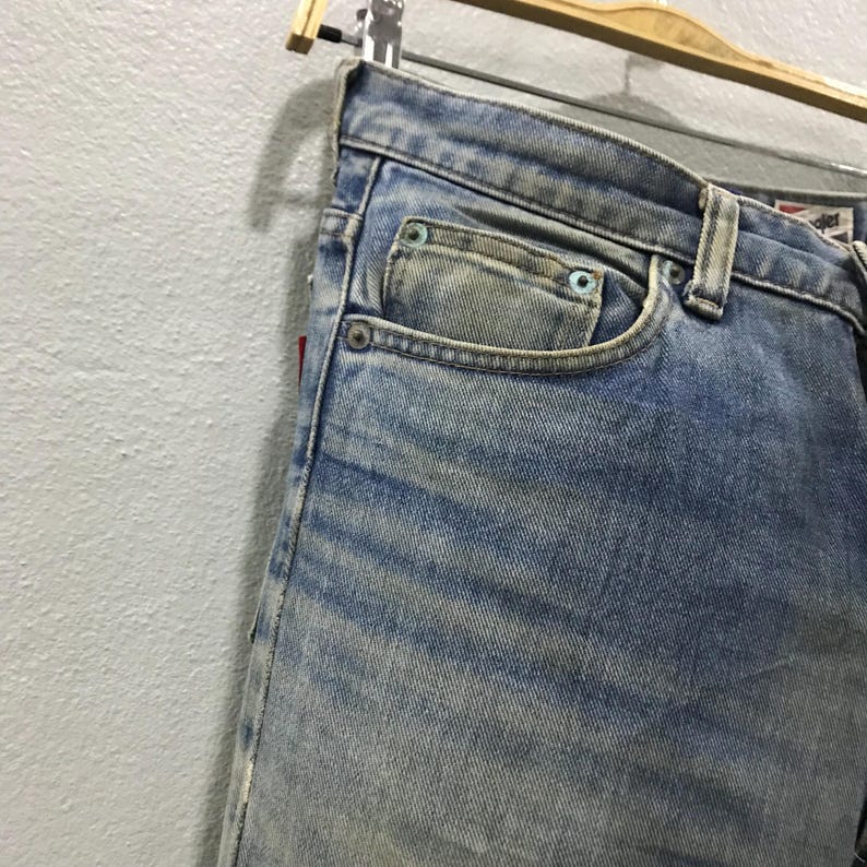 以下が含まれることがあります： 色あせたウォッシュ加工が施された、明るいブルーデニムのジーンズのクローズアップ。ジーンズは、小さなコインポケットを含む、クラシックな5ポケットデザインが特徴です。生地にはわずかな摩耗が見られ、側面に赤いタグが見えます。