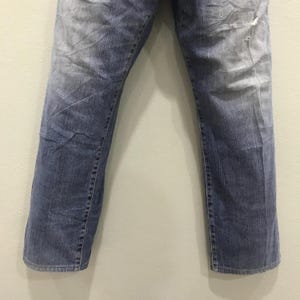 May include: Faded blue denim jeans with a distressed look. The jeans have a straight leg cut and show signs of wear and tear, including a small rip near the knee. The denim has a washed appearance with lighter areas.