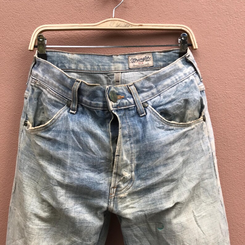 May include: Light blue Wrangler jeans with a faded wash, hanging on a wooden hanger. The jeans have a button closure and classic five-pocket design. The Wrangler label is visible at the waistband.