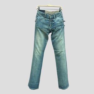 May include: Light blue denim jeans with unique design details, hanging on a hanger. The jeans feature a distinctive waistline and pocket design. The denim has a faded wash, giving it a vintage look. The jeans are straight-legged.