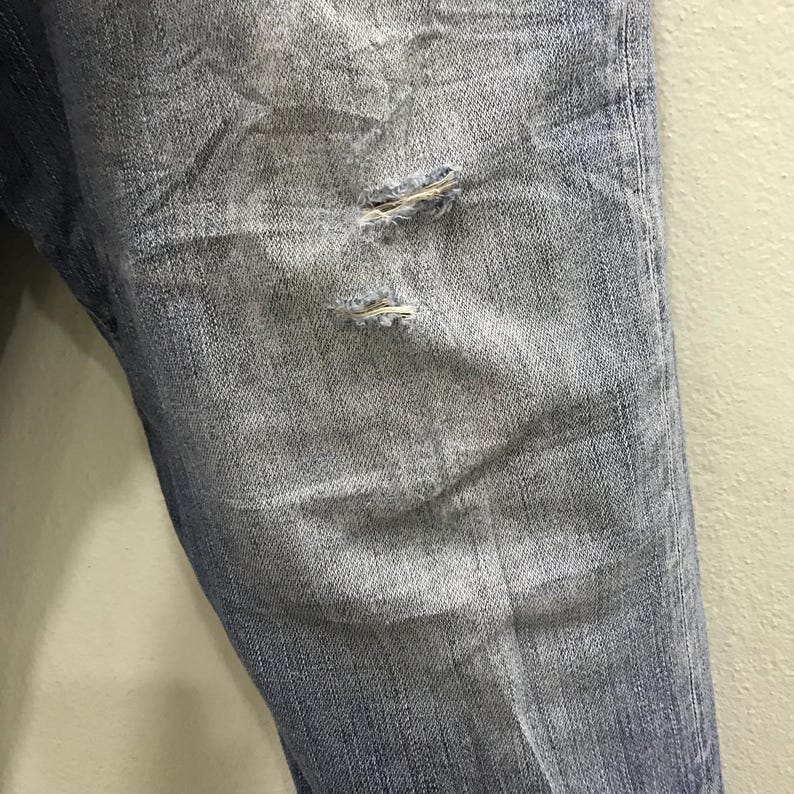 May include: Close-up of light blue denim jeans with distressed details. The jeans have two small, frayed holes near the knee area, adding a worn and casual aesthetic. The fabric shows subtle fading and texture.