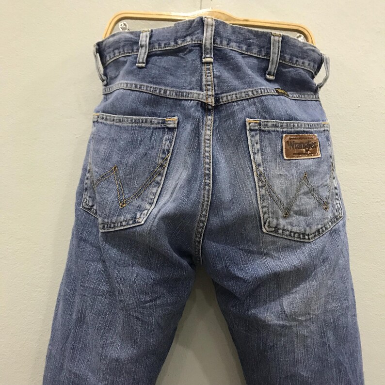 May include: Light blue denim jeans with the Wrangler logo on the back pocket. The jeans are hanging on a wooden hanger. The back pockets have a distinctive stitching pattern.