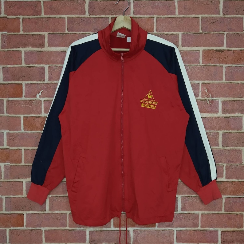 Puede incluir: Chaqueta de ch&aacute;ndal roja, negra y blanca con cierre de cremallera y un logotipo que dice "Le Coq Sportif Extreme".