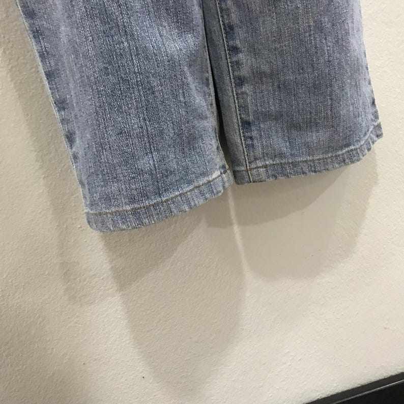 May include: Close-up of the hem of light blue denim jeans. The jeans are slightly faded and show signs of wear. The fabric is a medium weight, and the stitching is visible along the hem. The jeans are likely part of a casual wear collection.