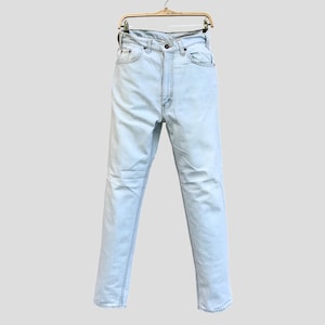 Könnte beinhalten: Hellblaue Jeans aus Denim, die an einem Holzbügel hängen. Die Jeans haben ein klassisches Fünf-Taschen-Design, einen Knopf- und Reißverschluss und eine gerade Beinpassform. Der Denim hat einen verblichenen, abgenutzten Look.