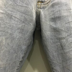 May include: Close-up of light blue denim jeans. The fabric shows wear and fading, with subtle texture and stitching details. The image focuses on the front of the jeans, highlighting the texture and colour variations.