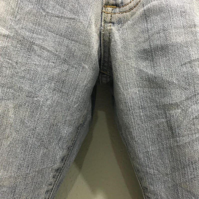 May include: Close-up of light blue denim jeans. The fabric shows wear and fading, with subtle texture and stitching details. The image focuses on the front of the jeans, highlighting the texture and colour variations.