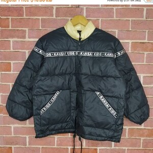 May include: Black puffer jacket with a white fleece lining and a zipper closure. The jacket has a band around the waist and the sleeves with the text "KANSAI KIDS" repeated.