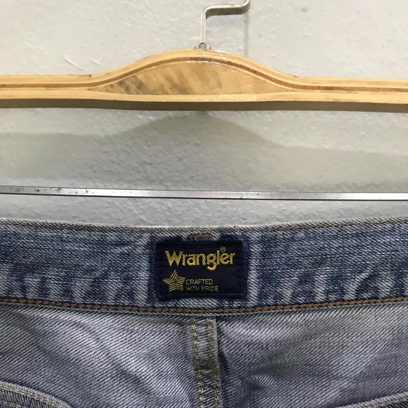May include: Close-up of a pair of blue denim jeans. The Wrangler brand label is visible, featuring gold lettering and a star emblem. The label reads "Wrangler Crafted With Pride."