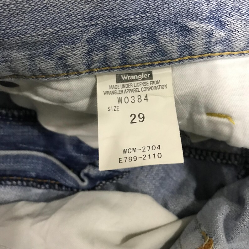 May include: Close-up of a light blue denim jeans label. The label reads "Wrangler" and includes the size "29". Additional text includes "MADE UNDER LICENSE FROM WRANGLER APPAREL CORPORATION", "W0384", "WCM-2704", and "E789-2110".