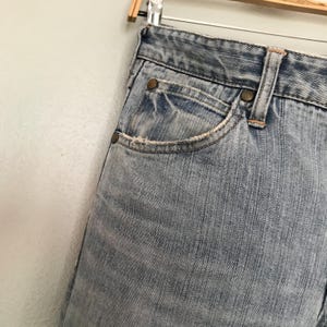K&ouml;nnte beinhalten: Nahaufnahme einer hellblauen Jeans mit verwaschener Optik. Die Jeans hat ein klassisches F&uuml;nf-Taschen-Design mit sichtbaren N&auml;hten und Metallnieten. Die Kanten der Taschen weisen leichte Abnutzungserscheinungen auf, was den Vintage-Look unterstreicht. Die Jeans h&auml;ngt an einem Holzb&uuml;gel.