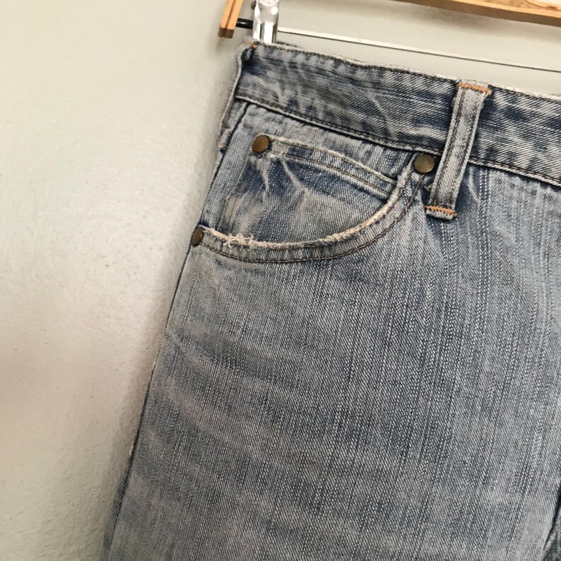 K&ouml;nnte beinhalten: Nahaufnahme einer hellblauen Jeans mit verwaschener Optik. Die Jeans hat ein klassisches F&uuml;nf-Taschen-Design mit sichtbaren N&auml;hten und Metallnieten. Die Kanten der Taschen weisen leichte Abnutzungserscheinungen auf, was den Vintage-Look unterstreicht. Die Jeans h&auml;ngt an einem Holzb&uuml;gel.