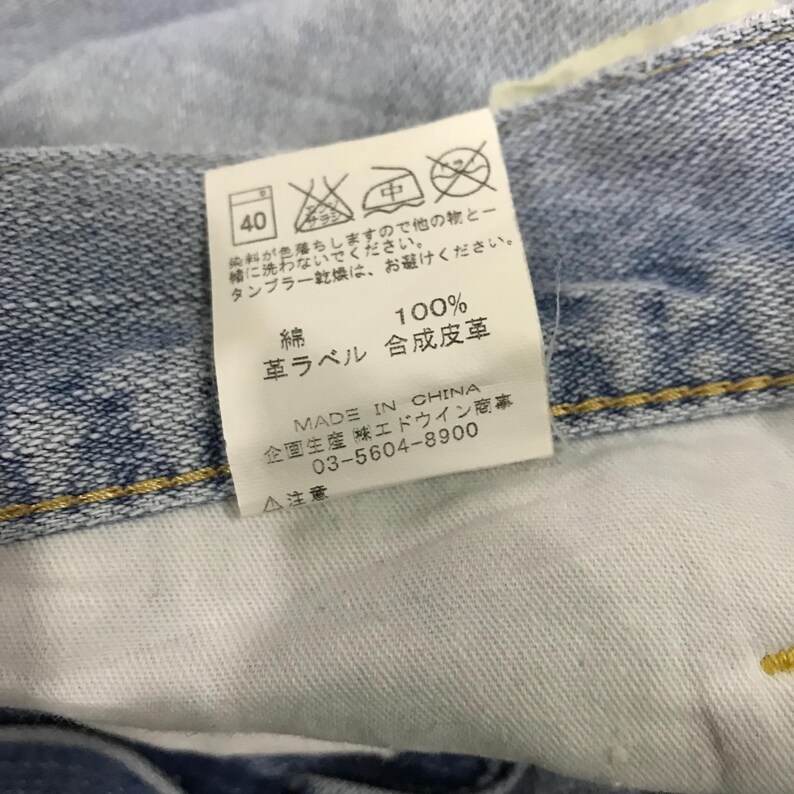 May include: A close-up shot of a light blue denim garment with a white label. The label features Japanese text and symbols, including washing instructions and fabric composition. The label reads "MADE IN CHINA".