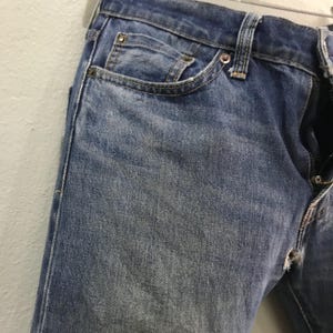 May include: Close-up of faded blue denim jeans. The jeans have a classic five-pocket design, with visible wear and tear, including a small tear near the zipper. The fabric shows a textured appearance, with lighter areas from wear.