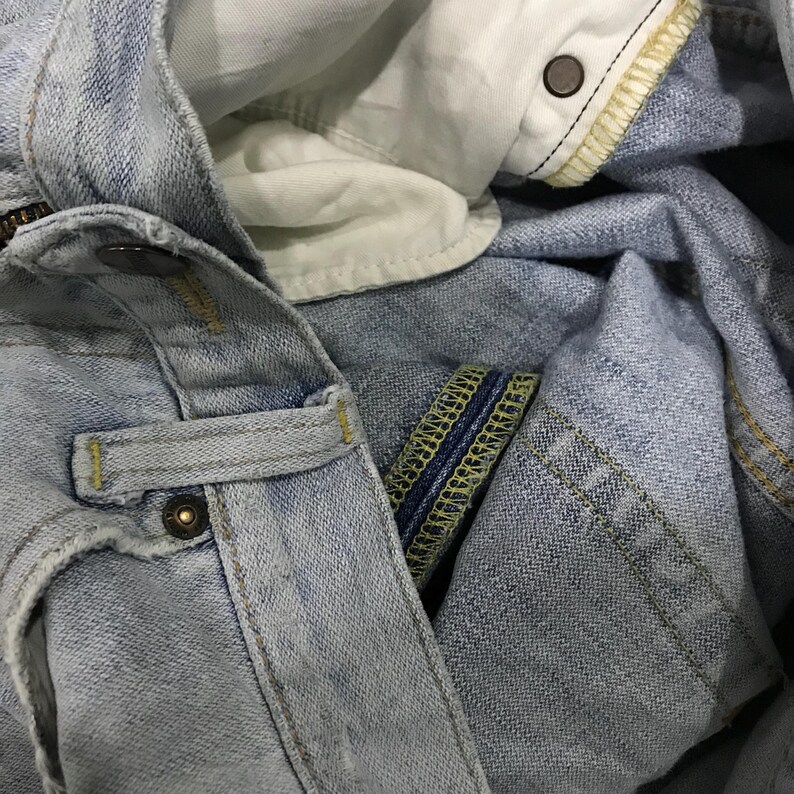 May include: Close-up of light blue denim jeans. Details include a button closure, belt loop, and the interior lining. Yellow stitching and a faded denim appearance are visible.