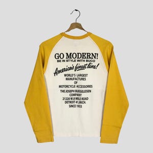 May include: A long-sleeved raglan shirt in cream and yellow, hanging on a hanger. The back of the shirt features the text "GO MODERN!" and "America's finest line!" along with information about a motorcycle accessories company.