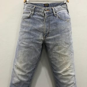 May include: Light blue Wrangler jeans, displayed on a hanger. The jeans have a faded appearance with lighter patches on the thighs and knees. The Wrangler logo is visible on the waistband. The jeans are a classic, straight-leg style.