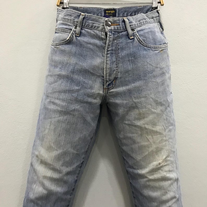 May include: Light blue Wrangler jeans, displayed on a hanger. The jeans have a faded appearance with lighter patches on the thighs and knees. The Wrangler logo is visible on the waistband. The jeans are a classic, straight-leg style.