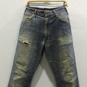 以下が含まれることがあります： 色あせた青色のデニムジーンズ。左脚に大きな穴があり、ダメージ加工が施されています。ジーンズはクラシックな5ポケットデザインで、木製のハンガーに吊るされています。