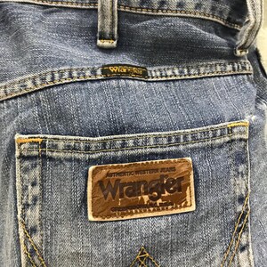 May include: Close-up of the back pocket of blue denim Wrangler jeans. The pocket features a brown leather patch with the Wrangler logo and the words "Authentic Western Jeans". The jeans have yellow stitching.