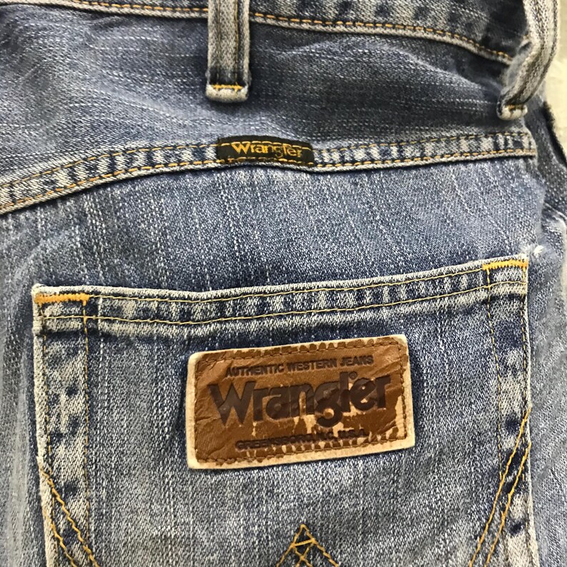 May include: Close-up of the back pocket of blue denim Wrangler jeans. The pocket features a brown leather patch with the Wrangler logo and the words "Authentic Western Jeans". The jeans have yellow stitching.